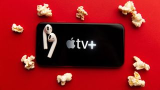 Apple TV Plus 로고가 있는 iPhone, 상단에 AirPods 한 쌍, 팝콘 조각이 있는 빨간색 배경 위에 놓여 있음