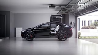 흰색 차고에 가정용 충전기 옆에 앉아 있는 검은색 Tesla Model Y